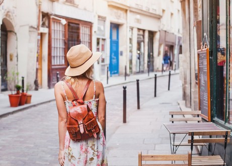 A woman wearing a small leather backpack walks down a narrow old city street with a brick road to her left and an outdoor cafe on her right.